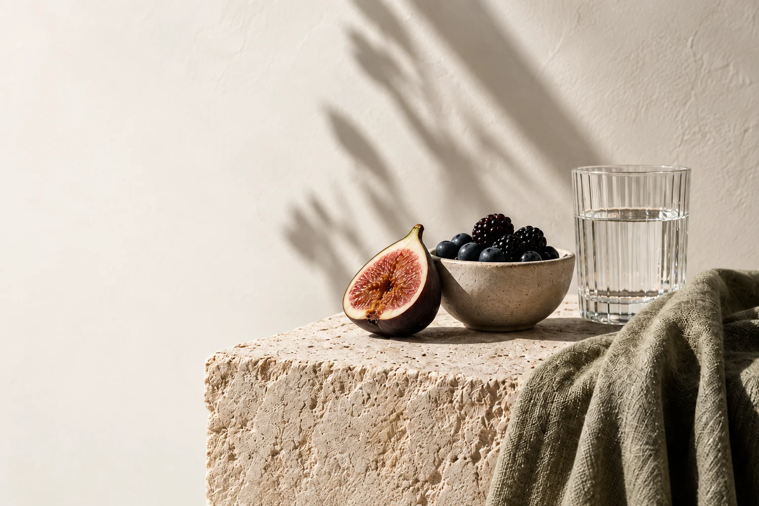 Fresh figs, berries and a water glass on sunlit limestone with sage linen